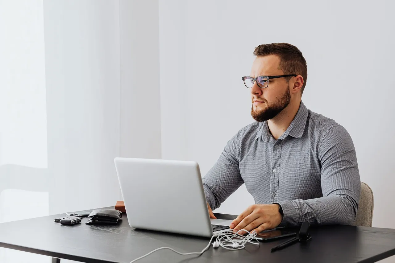 Bearded man with eyeglasses working on a laptop in a minimalist office setting.