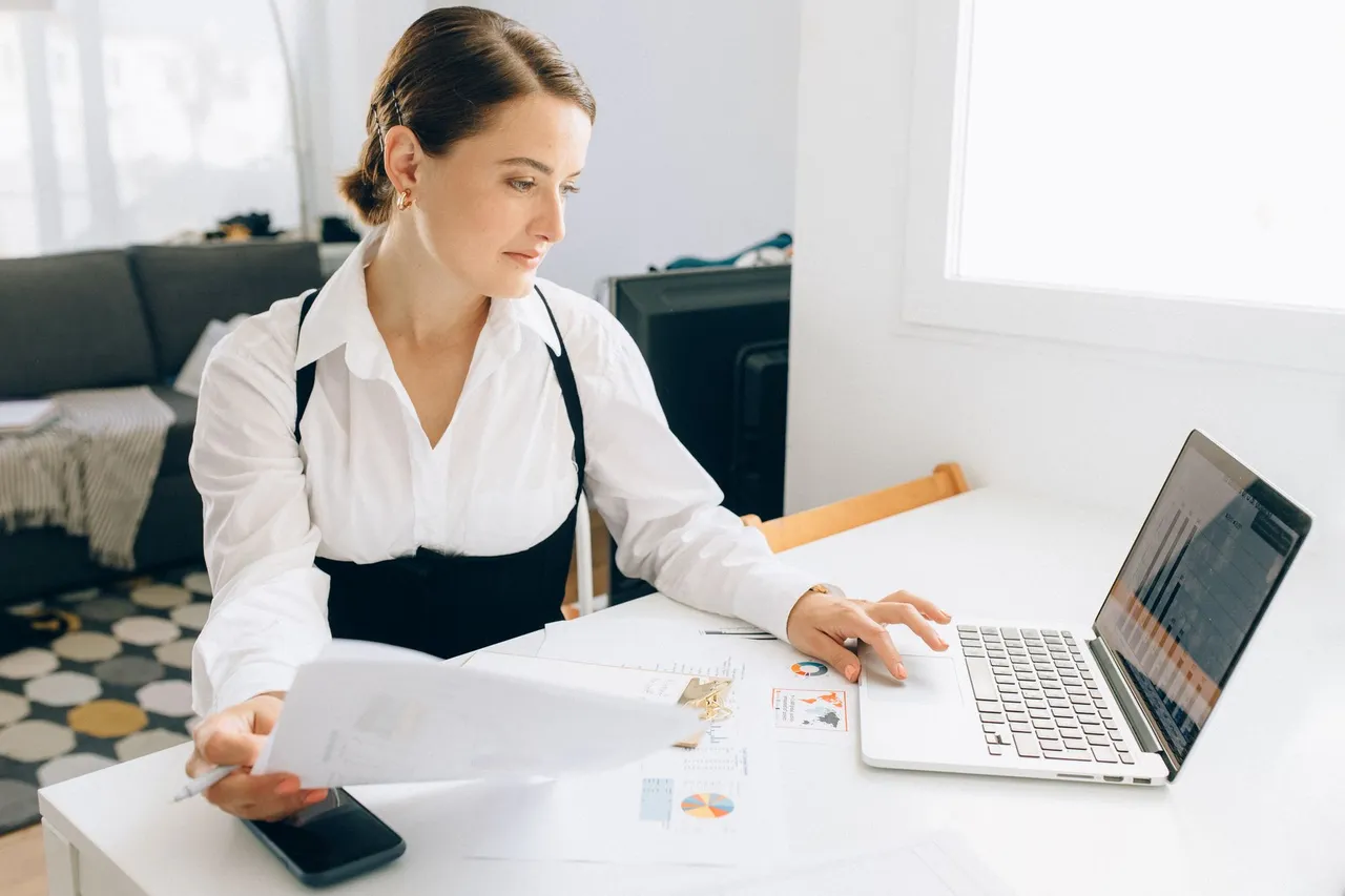Woman at desk reviewing business reports on laptop, holding papers in a modern office setting.
